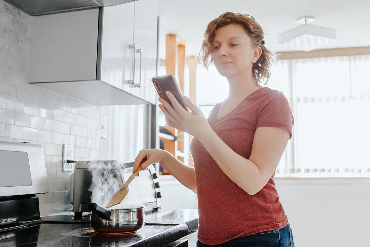 Young woman in kitchen watches instructional video on a smartphone whilst stirring a saucepan.
