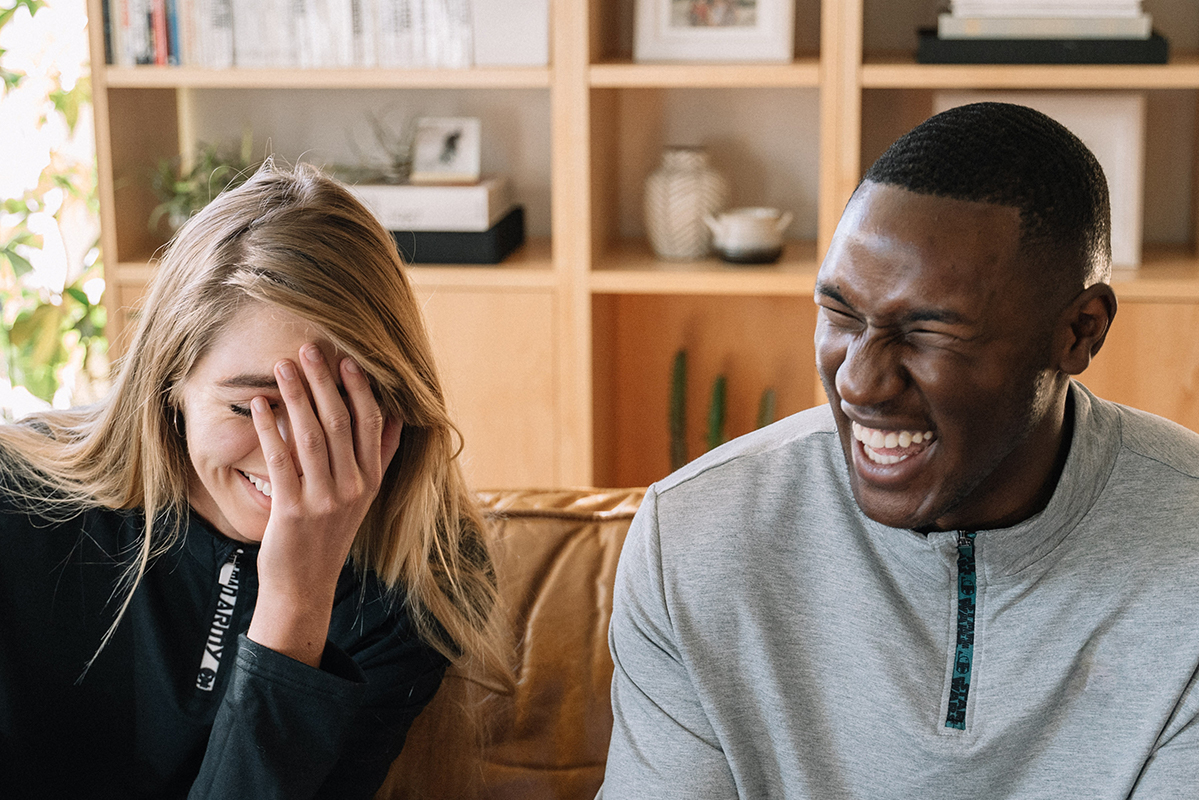 White woman and black man sit in a living room and are laughing hard at a joke.