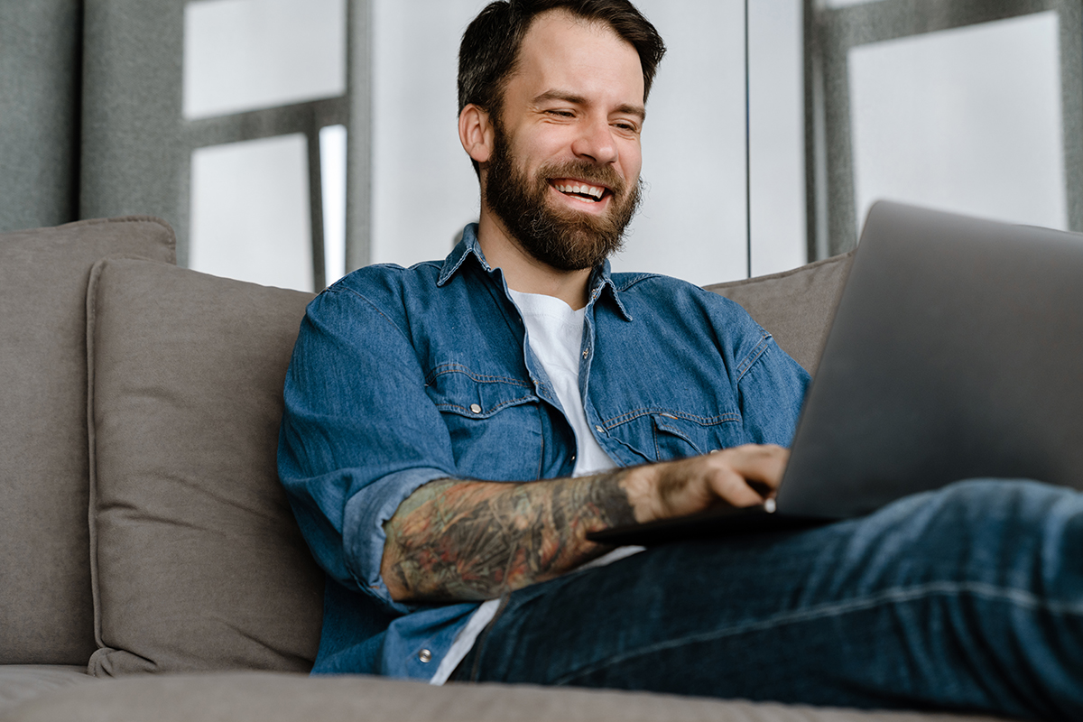 man met baard zit in zetel met laptop op schoot, is enthousiast en lacht terwijl hij een e-learning volgt rond veiligheid op het werk